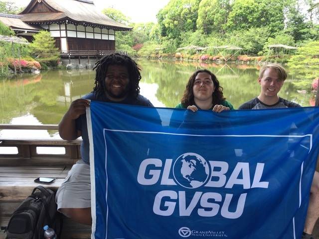 Three students in front of a pond holding a GVSU flag
